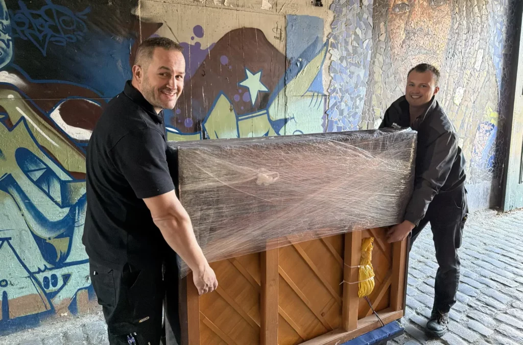 Twee verhuizers van Hoekema Verhuizingen rijden een ingepakte piano op een pianowagen door een steeg